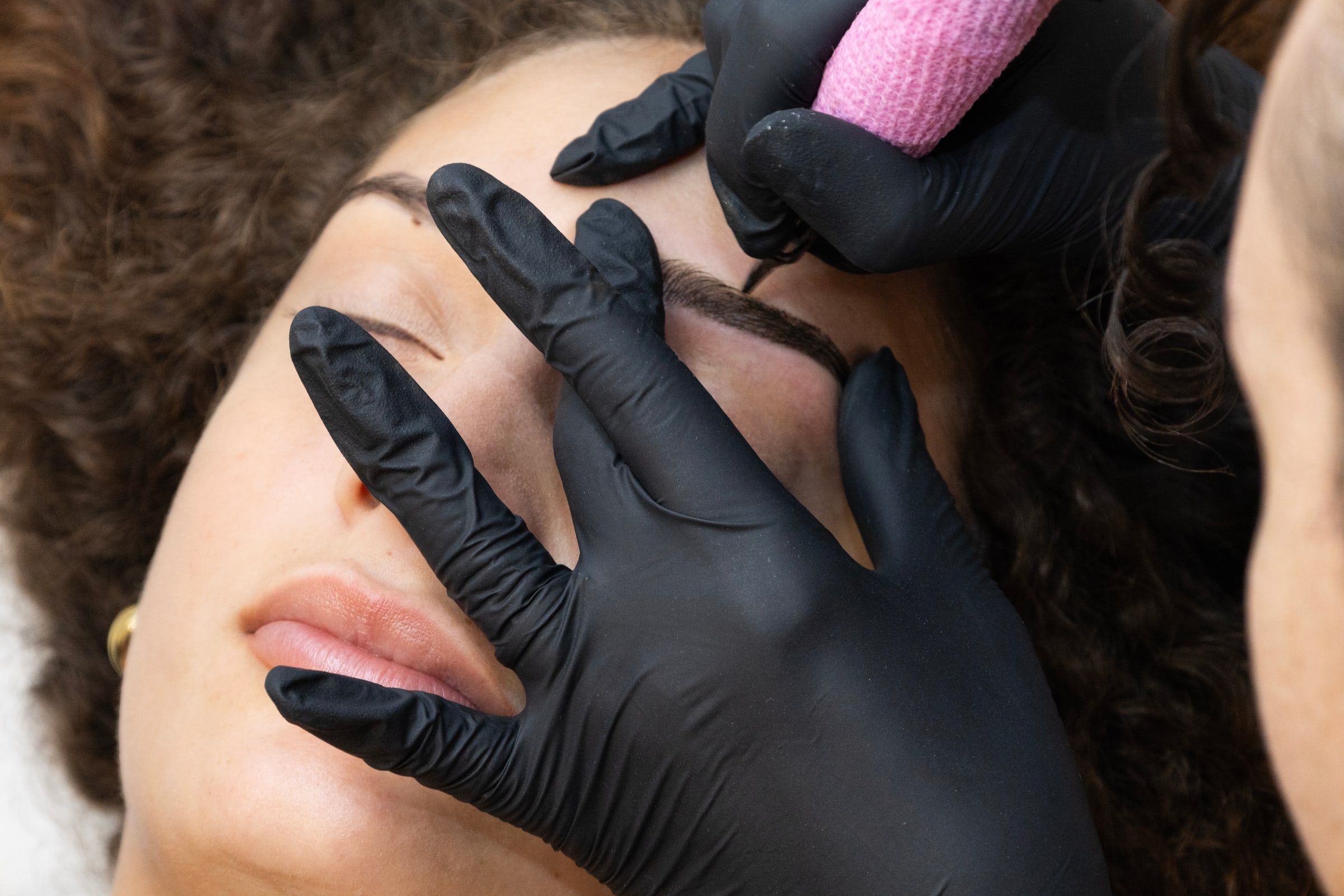 Le mani con i guanti neri tengono ferma la sopracciglia mentre il dermografo deposita il pigmento